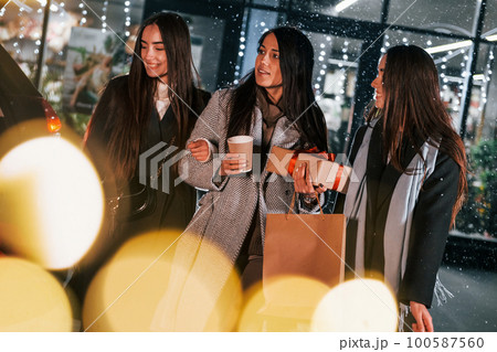 Cups of drink in hands. Three cheerful women spends Christmas holidays together outdoors. Conception of new year 100587560