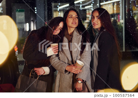 Cups of drink in hands. Three cheerful women spends Christmas holidays together outdoors. Conception of new year 100587564