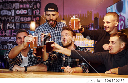 Cheering by knocking beer glasses. Group of people together indoors in the pub have fun at weekend time 100590282