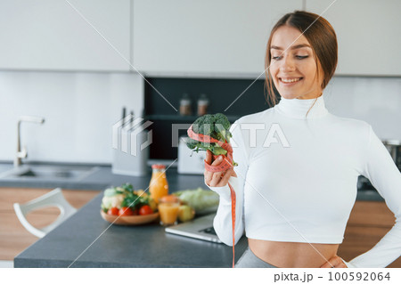 Poses for a camera with vegetables. Young european woman is indoors at kitchen indoors with healthy food 100592064