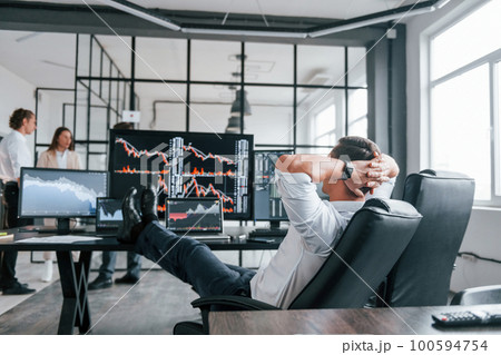 Man sits on chair and takes break. Team of stockbrokers works in modern office with many display screens 100594754