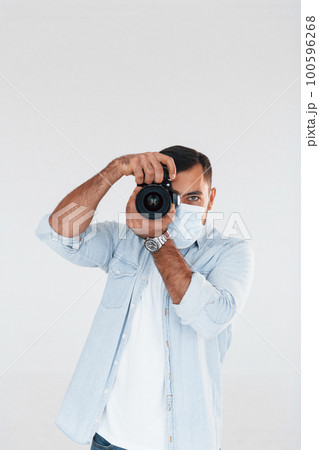 With professional camera. Young handsome man standing indoors against white background With professional camera. Young handsome man standing indoors against white background 100596268