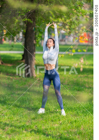 Young woman stretching body - warming up before running or working out. Female runner stretching arms before running at morning. Vertical photo. 100607408