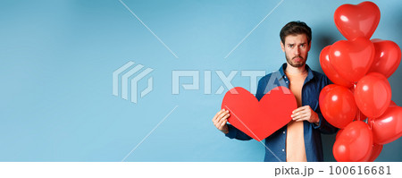 Sad man looking heartbroken and lonely, holding paper red heart and standing near balloons over blue background 100616681