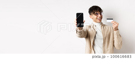 E-commerce. Happy young man looking satisfied at plastic credit card, showing smartphone screen to boast, standing on white background E-commerce. Happy young man looking satisfied at plastic credit card, showing smartphone screen to boast, standing on white background 100616683