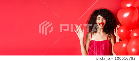 Excited stylish woman with curly hair, wearing dress, raising hands up and laughing happy, standing near heart balloons, white background 100616783