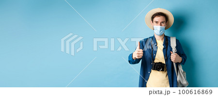 Covid-19 and summer holidays concept. Smiling guy travelling in medical mask and straw hat, backpacking and showing thumbs up, approve and praise travel agency, blue background Covid-19 and summer holidays concept. Smiling guy travelling in medical mask and straw hat, backpacking and showing thumbs up, approve and praise travel agency, blue background 100616869