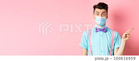 Coronavirus, healthcare and quarantine concept. Man looking surprised in medical mask, asking question and pointing right, curious about promo offer, standing on pink background Coronavirus, healthcare and quarantine concept. Man looking surprised in medical mask, asking question and pointing right, curious about promo offer, standing on pink background 100616959