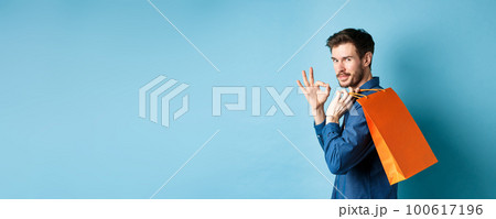 Handsome young man holding shopping bag on shoulder, turn around to look at camera and show okay gesture, recommending shop, blue background 100617196