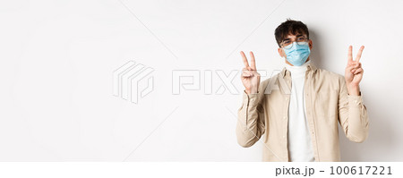 Covid-19, health and real people concept. Cute happy guy in medical mask staying positive, showing peace signs and smiling, standing on white background Covid-19, health and real people concept. Cute happy guy in medical mask staying positive, showing peace signs and smiling, standing on white background 100617221