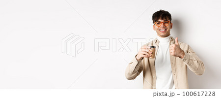Real people. Cheerful smiling guy in glasses drinking coffee and showing thumb up, recommending cafe, standing on white background Real people. Cheerful smiling guy in glasses drinking coffee and showing thumb up, recommending cafe, standing on white background 100617228