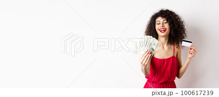 Shopping. Rich successful woman with curly hair and red dress, holding plastic credit card and looking pleased at dollar bills, white background Shopping. Rich successful woman with curly hair and red dress, holding plastic credit card and looking pleased at dollar bills, white background 100617239