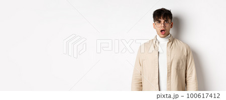 Portrait of shocked young man in glasses drop jaw, open mouth and stare amazed at camera, looking at something impressive, standing on white background 100617412