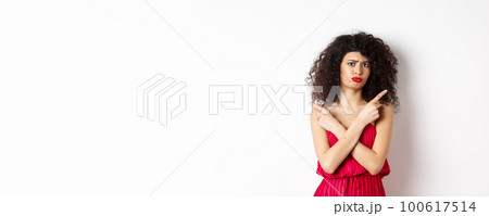 Doubtful and sad young woman in red dress, pointing fingers sideways and sulking, cant decide between products, need help with choice, standing over white background 100617514
