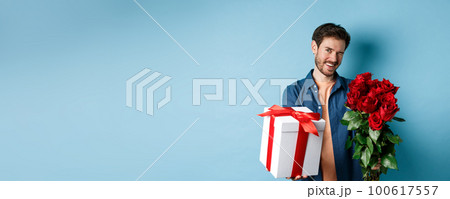Love and Valentines day concept. Happy guy winking and smiling at camera, presenting a gift and bouquet of flowers to girlfriend, standing over blue background Love and Valentines day concept. Happy guy winking and smiling at camera, presenting a gift and bouquet of flowers to girlfriend, standing over blue background 100617557
