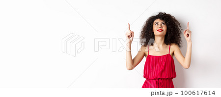 Dreamy young woman in trendy red dress, looking up and pointing at logo, smiling happy, standing over white background Dreamy young woman in trendy red dress, looking up and pointing at logo, smiling happy, standing over white background 100617614