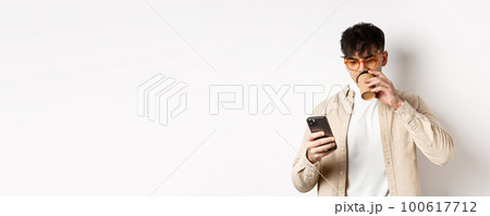 Real people. Young man reading smartphone screen and drinking coffee, looking at phone, standing on white background Real people. Young man reading smartphone screen and drinking coffee, looking at phone, standing on white background 100617712