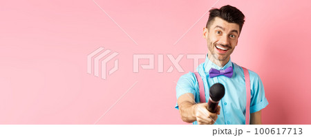 Smiling guy with moustache and bow-tie asking for comments or interview, stretch out hand with microphone, standing on pink background Smiling guy with moustache and bow-tie asking for comments or interview, stretch out hand with microphone, standing on pink background 100617713