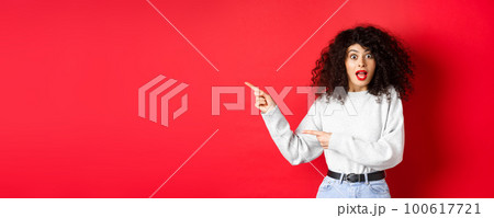 Excited caucasian woman in sweatshirt, checking out banner, pointing fingers left at empty space, showing advertisement, red background Excited caucasian woman in sweatshirt, checking out banner, pointing fingers left at empty space, showing advertisement, red background 100617721