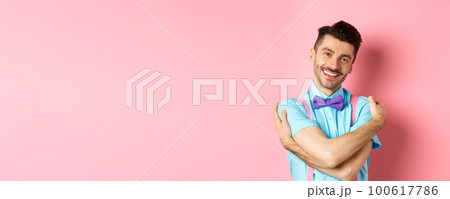 Cheerful young man smiling and hugging himself, wrap hands around own body and looking happy, standing over pink background 100617786