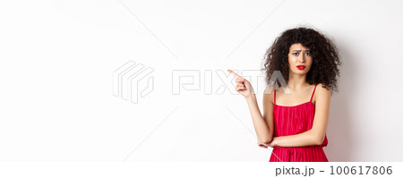 Arrogant young woman with curly hair, wearing red dress, frowning and complaining, pointing finger left at promo, standing over white background 100617806