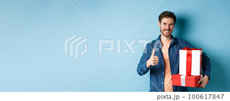 Smiling man holding romantic gifts and showing thumbs-up, celebrating Valentines day, buying presents for lover, standing over blue background 100617847