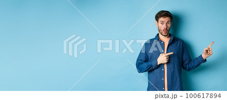 Surprised young man checking out promo, pointing fingers right at logo and saying wow, standing on blue background Surprised young man checking out promo, pointing fingers right at logo and saying wow, standing on blue background 100617894