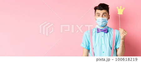Covid, pandemic and quarantine concept. Cute and silly guy in medical mask looking surprised, holding small crown for party, standing over pink background Covid, pandemic and quarantine concept. Cute and silly guy in medical mask looking surprised, holding small crown for party, standing over pink background 100618219