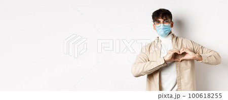Health, covid and quarantine concept. Romantic young man in sterile medical mask showing heart gesture on chest, say I love you, standing on white background 100618255