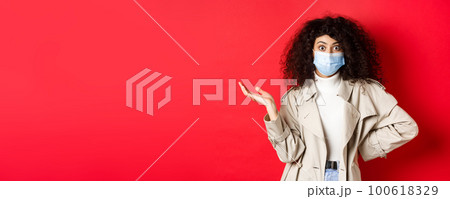 Covid-19, social distancing and quarantine concept. Shocked and confused european woman with curly hair, wearing medical mask from coronavirus, raise hand up puzzled, red background Covid-19, social distancing and quarantine concept. Shocked and confused european woman with curly hair, wearing medical mask from coronavirus, raise hand up puzzled, red background 100618329