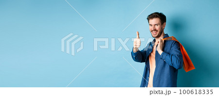 Handsome young guy with shopping bag showing thumbs up and smiling, praising good promotion offer, standing on blue background Handsome young guy with shopping bag showing thumbs up and smiling, praising good promotion offer, standing on blue background 100618335
