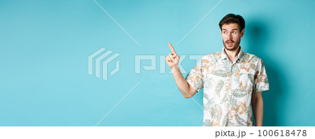 Intrigued man in hawaiian shirt, looking and pointing left at something interesting, blue background. Concept of tourism and vacation Intrigued man in hawaiian shirt, looking and pointing left at something interesting, blue background. Concept of tourism and vacation 100618478
