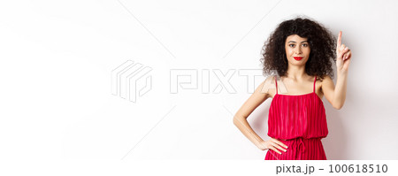 Smiling caucasian woman with curly hair, wearing red dress, showing rule number one gesture, raising finger and looking confident at camera, white background Smiling caucasian woman with curly hair, wearing red dress, showing rule number one gesture, raising finger and looking confident at camera, white background 100618510