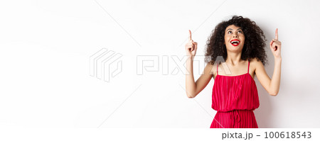 Excited caucasian woman with curly hair, red lips, looking and pointing up with amazed smile, checking out promo offer, white background 100618543