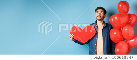 Valentines day and love concept. Man dreaming of soulmate, holding big red heart cutout and balloons, standing over blue background 100618547