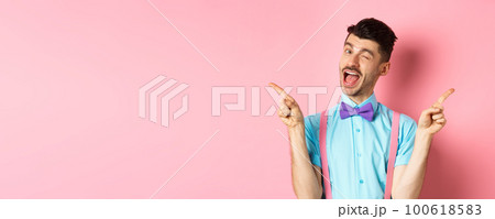 Cheerful young man smiling and winking, showing two sides, pointing fingers sideways and recommending promo offers on pink background 100618583