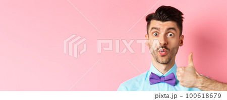 Close-up of funny guy with moustache, say wow and showing thumb up in approval, checking out something cool, recommending product, standing on pink background 100618679