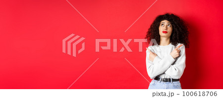 Indecisive female model with red lips and curly hair, looking up troubled with making choice, pointing fingers sideways, choosing between two products, studio background 100618706