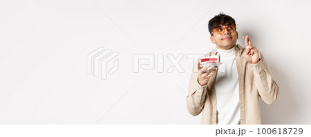 Cute hopeful guy in glasses making birthday wish, holding cake and fingers crossed, looking up and praying, hope dream come true, standing on white background Cute hopeful guy in glasses making birthday wish, holding cake and fingers crossed, looking up and praying, hope dream come true, standing on white background 100618729