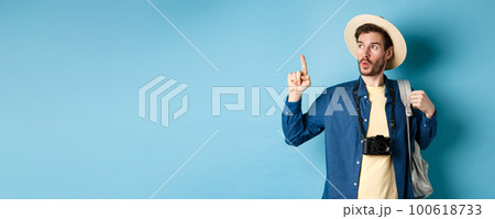 Handsome tourist looking curious and pointing up, showing interesting sightseeing on summer vacation, wearing straw hat and holding backpack with camera, blue background 100618733