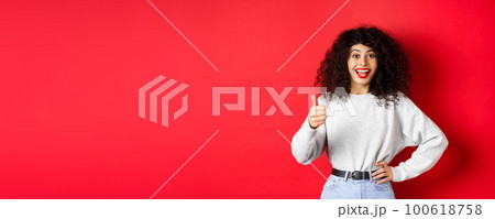 Happy young woman with curly hair praising good work, say well done and show thumb up gesture, approve and praise you, standing on red background Happy young woman with curly hair praising good work, say well done and show thumb up gesture, approve and praise you, standing on red background 100618758
