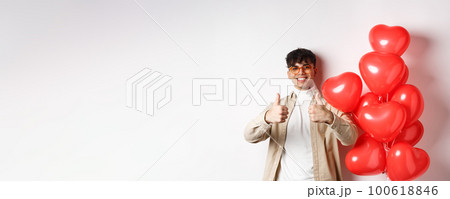 Valentines day and romance concept. Stylish modern man in sunglasses showing thumbs up while standing near heart balloons, saying yes, enjoy date with lover Valentines day and romance concept. Stylish modern man in sunglasses showing thumbs up while standing near heart balloons, saying yes, enjoy date with lover 100618846
