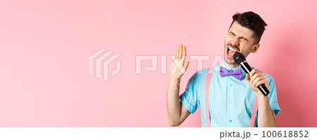 Party and festive events concept. Funny guy performs a song, singing karaoke in microphone with carefree face, standing in bow-tie and suspenders on pink background 100618852