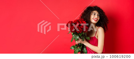 Tender young woman in elegant red dress, holding romantic bouquet of red roses and looking right, smiling dreamy, thinking about lover on Valentines day, studio background 100619200