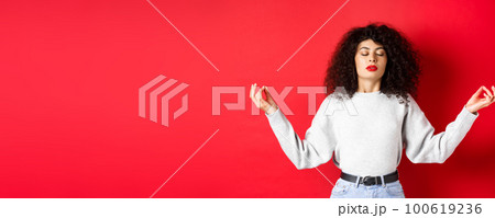 Woman in casual clothes trying to calm down with meditation, close eyes and practice yoga, holding hands sideways with zen gesture, standing on red background 100619236