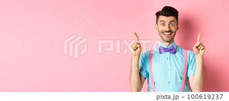 Portrait of cheerful young guy with moustache and bristle pointing fingers up, showing top logo, smiling happy at camera, standing on pink background 100619237