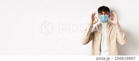 Covid-19, health and real people concept. Handsome guy in glasses and medical mask showing okay gesture in approval, recommend use preventive measures from corona, white background Covid-19, health and real people concept. Handsome guy in glasses and medical mask showing okay gesture in approval, recommend use preventive measures from corona, white background 100619897