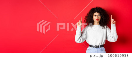 Grumpy young woman with curly hair frowning and grimacing unsatisfied, pointing fingers up at something bad, complaining on company, red background Grumpy young woman with curly hair frowning and grimacing unsatisfied, pointing fingers up at something bad, complaining on company, red background 100619953
