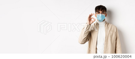 Covid-19, health and real people concept. Natural guy in glasses and medical mask show Ok sign, agree or approve something, standing on white background Covid-19, health and real people concept. Natural guy in glasses and medical mask show Ok sign, agree or approve something, standing on white background 100620004