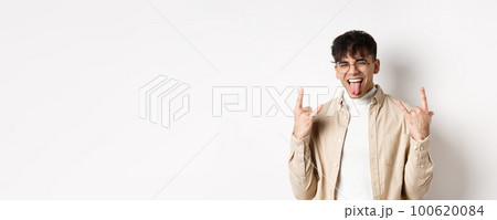 Happy handsome guy showing tongue and rock-on gesture, having fun and feeling upbeat, enjoying something good, standing on white background 100620084